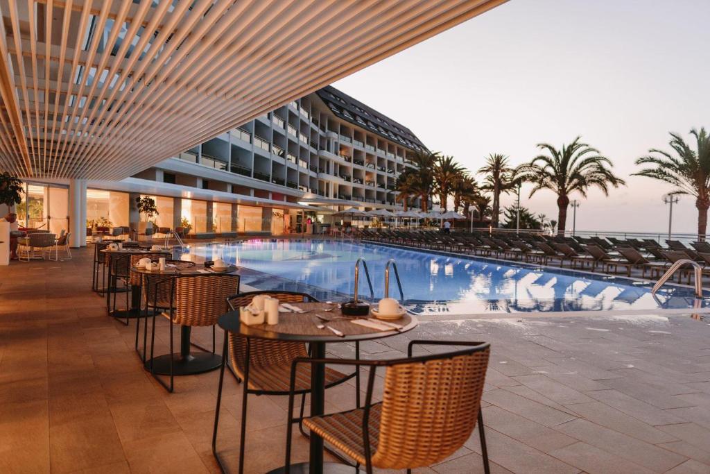 Mūsdienīgs viesnīcas restorāns ar terasi pie baseina, kurā redzami galdi ar ēdienu un dzērieniem, kā arī palmām un viesnīcas ēka fona. Ideāls vieta relaksācijai pie ūdens.