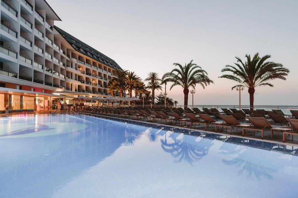 Moderns viesnīcas komplekss ar baseinu un palmu rindām, kas atspoguļojas ūdenī, radot relaksējošu atmosfēru pie jūras. Ideāla vieta atpūtai un brīvdienām.