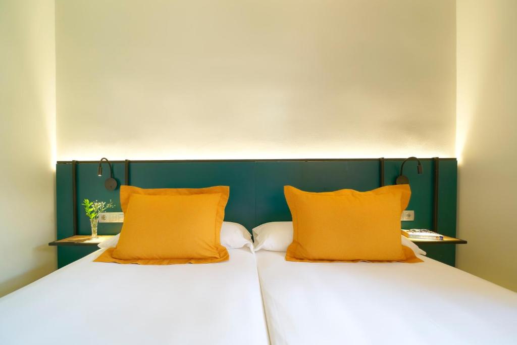 Mūsdienīgs guļamistabas interjers ar diviem gultas veļas komplektiem un spilveniem dzeltenā krāsā. Uz galda atrodas ziedi un gaismas avots, radot siltu atmosfēru.