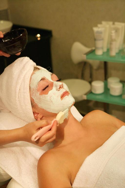 Sejas maskas uzklāšana spa procedūrā, kur sieviete relaksējas ar baltu masku uz sejas. Fons veidots no kosmētikas produktiem un relaksējošas vides.