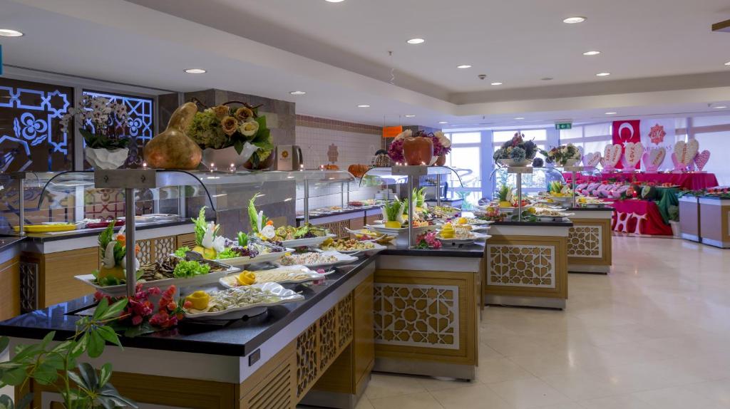 Bagātīgs bufetes galds ar dažādiem ēdieniem un rotājumiem, kas izkārtoti elegantā restorānā. Gaismas un krāsainas ziedu kompozīcijas piešķir atmosfēru šim gardēžu svētkam.