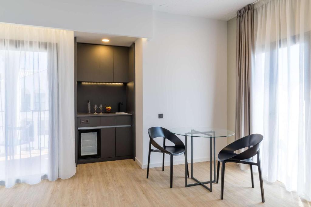 Mūsdienīgs viesistabas interjers ar stikla galdu un divām melnām krēsliem, blakus ir mini virtuve ar moderniem skapjiem un logiem ar gaisīgiem aizkariem.