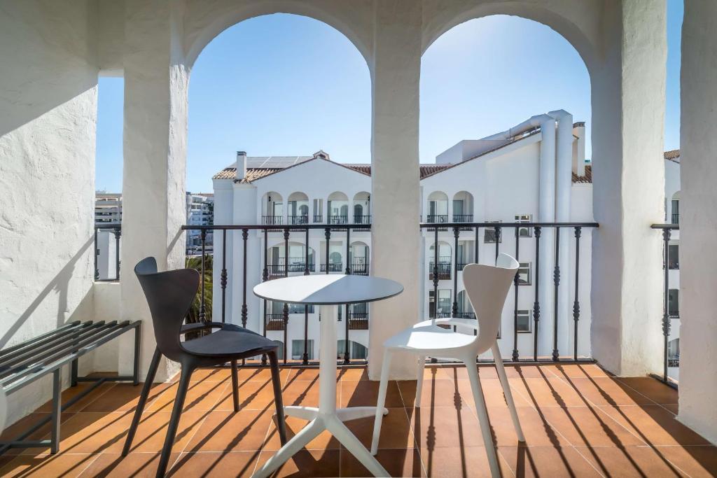 Mūsdienīgs balkons ar divām krāsainām krēslām un apaļu galdu, kas piedāvā skatu uz baltām ēkām un saulainu debesi. Ideāla vieta relaksācijai un baudīšanai.