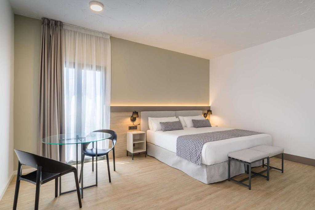Moderns viesnīcas numurs ar lielu gultu, galdu un krēsliem, izceļoties ar minimālistisku dizainu un dabīgu apgaismojumu.