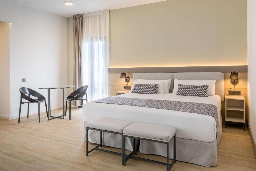 Mūsdienīga viesnīcas istaba ar divguļamo gultu, vienkāršām mēbelēm un elegantām apgaismojuma detaļām, kā arī galds un krēsli, kas piedāvā komfortu un funkcionālu dizainu.