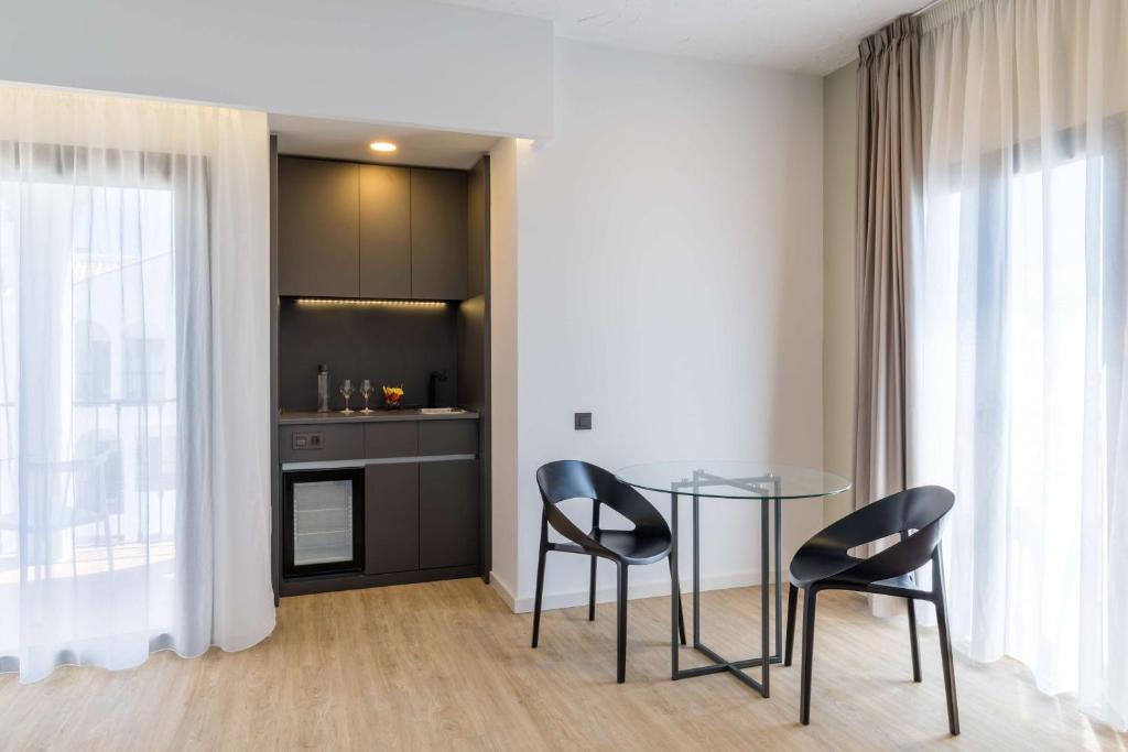 Mūsdienīga viesnīcas istaba ar gaišu interjeru, stikla galdu un melnām krēslām. Fons ietver nelielu virtuvi ar modernu aprīkojumu un logiem ar gaisīgām aizkariem.