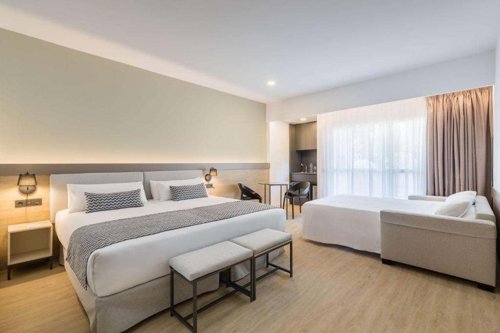 Mūsdienīga viesnīcas numura iekšskats ar diviem gultām, dīvānu un virtuves stūri. Telpa ir gaiša un plaša, ar lieliem logiem un neitrālām krāsām.