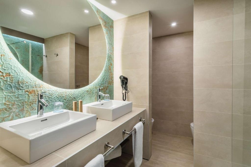 Mūsdienīgs vannas istabas interjers ar divām izlietnēm, elegantu spoguli un modernām flīzēm, kas piešķir telpai gaišu un svaigu izskatu.