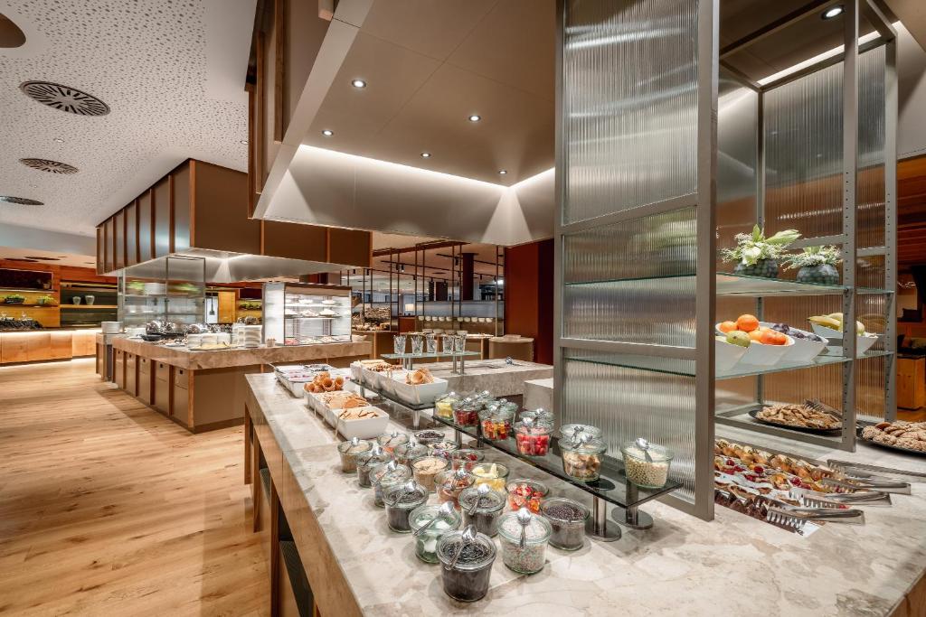 Mūsdienīgs bufetes stils ar dažādiem ēdieniem, tostarp augļiem un konditorejas izstrādājumiem, izkārtots eleganti un pievilcīgi. Gaismas un koka elementi rada siltu un viesmīlīgu atmosfēru.