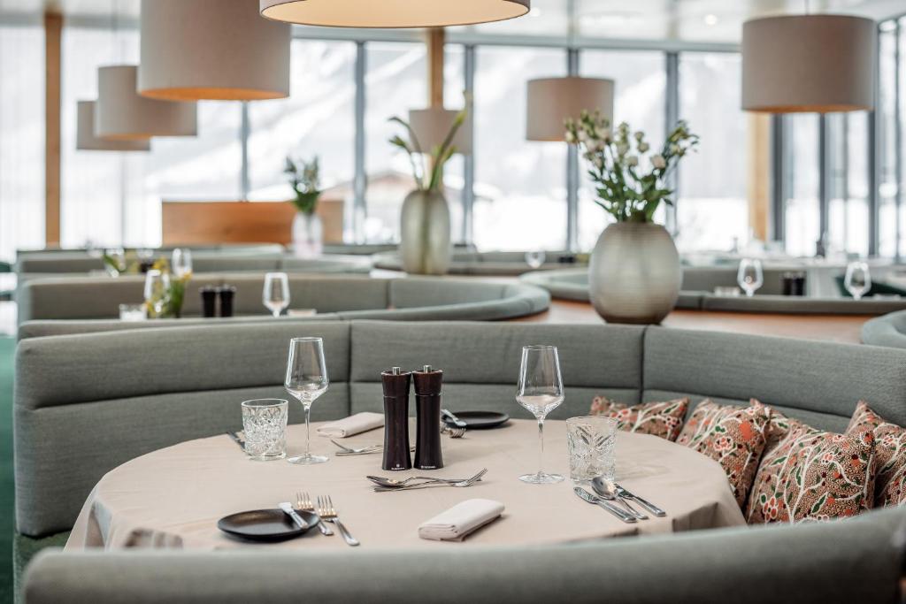 Elegants restorāns ar gaišu interjeru, modernām lampām un grezniem galdiem, kas sagatavoti viesu uzņemšanai. Skats uz kalniem caur lieliem logiem rada patīkamu atmosfēru.