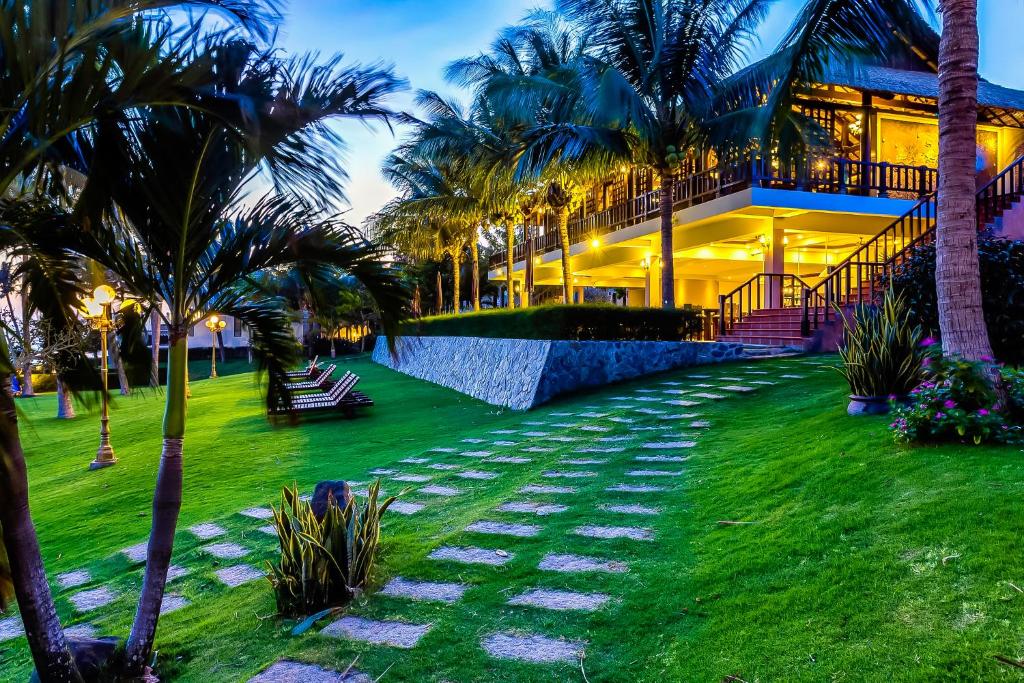 Viesmīlīgas viesnīcas dārzs ar zālienu un palmām, kas apgaismots vakara gaismā, radot relaksējošu atmosfēru. Celtnes arhitektūra un ainava izceļ tropisku brīvdienu noskaņu.