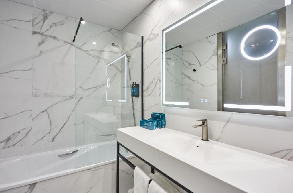 Mūsdienīgs vannas istabas interjers ar marmora efektu sienām, stikla dušas kabīni un izgaismoto spoguli. Balta izlietne ar modernu jaucējkrānu papildina eleganto dizainu.