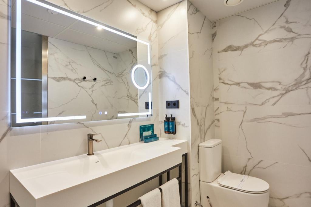 Moderns vannas istabas interjers ar marmora sienām, izgaismota spoguli un minimālistisku izlietni. Uz plaukta ir higiēnas līdzekļu komplekts.