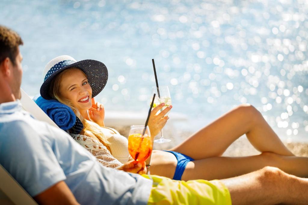 Pāris atpūšas pludmalē, baudot kokteiļus saulainā dienā. Viņi ir apģērbušies pludmales apģērbā, un fons ir skaists ūdens ar mirdzošām atspulgām.