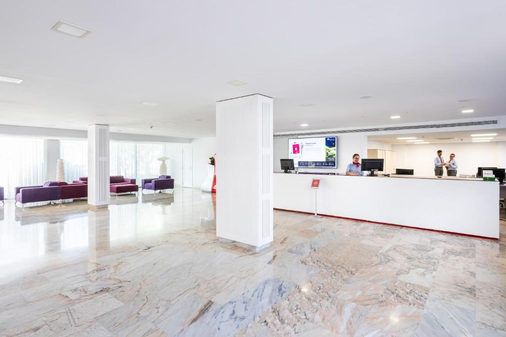 Mūsdienīga viesnīcas recepcija ar plašu, gaišu telpu un elegantām mēbelēm. Redzami darbinieki, kas sniedz pakalpojumus viesiem.