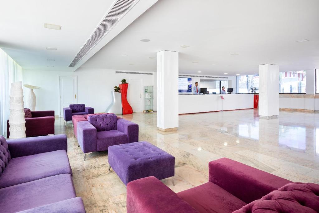 Mūsdienīga viesnīcas recepcija ar elegantiem violetiem dīvāniem un marmora grīdu, kas nodrošina plašu un gaisīgu atmosfēru. Recepcijas darbinieki apkalpo viesus pie baltas reģistratūras.