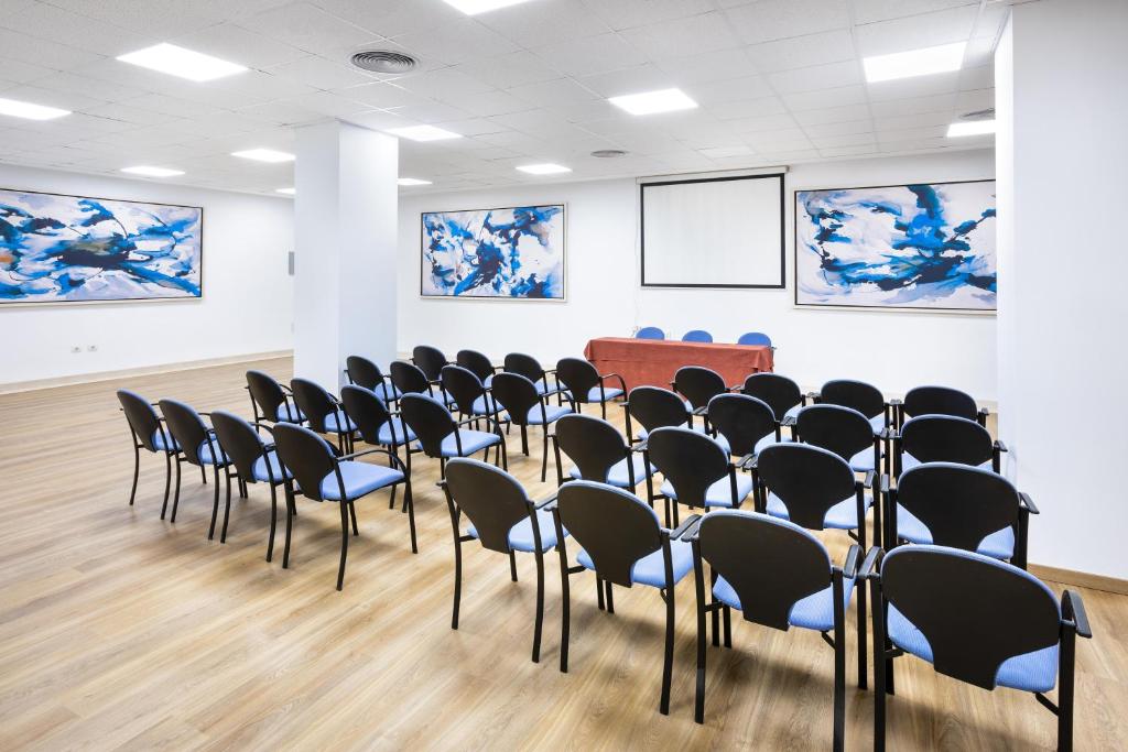 Konferenču telpa ar melnām krēslām un zilu polsterējumu, priekšā sarkanā galds un divi mākslas darbi ar zilu un baltu krāsu. Ideāla vide sanāksmēm un prezentācijām.