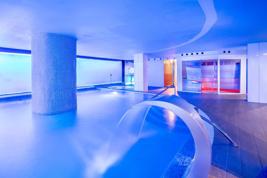 Mūsdienīgs spa ar zilu apgaismojumu un relaksējošu ūdens vidi, kurā redzama baseina un saunu zona.
