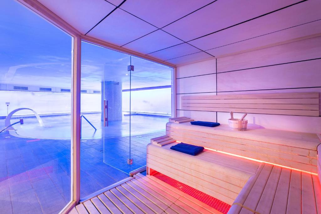 Mūsdienīga sauna ar koka soliņiem un zilu apgaismojumu, kas rada relaksējošu atmosfēru. Logu aizmugurē redzams baseins.
