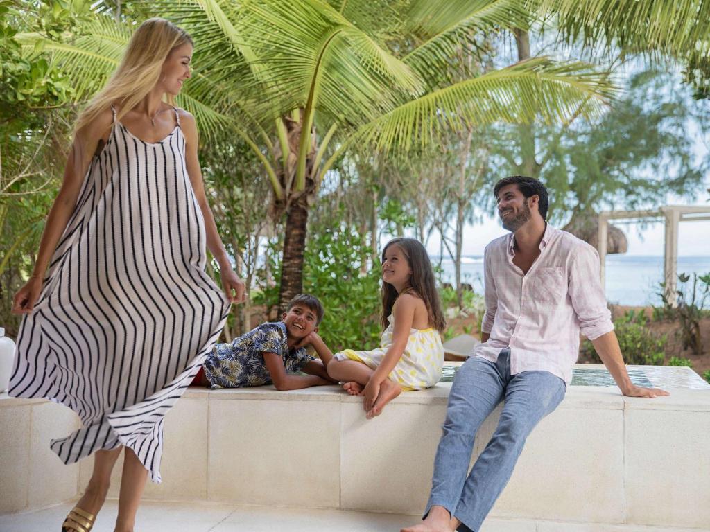 Ģimenes brīvdienu mirklis pie jūras, kur mamma, tērpta svārkos ar svītrām, vijīgi staigā, kamēr divi bērni un tēvs sēž uz akmens apmales un priecīgi sarunājas. Daba apkārt ir zaļa un saulaina.