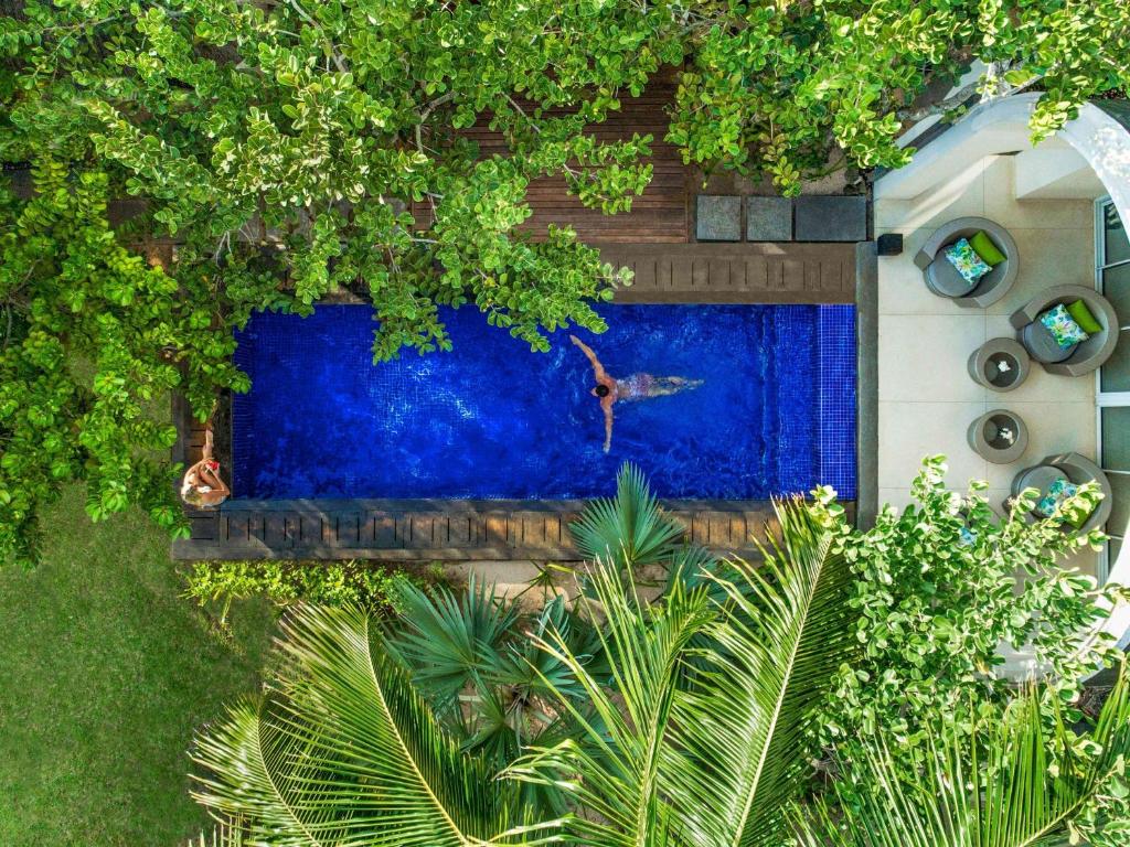 Peldbaseins ar zilu ūdeni, ko ieskauj zaļas lapas un tropu augi, un cilvēks peld ar izstieptām rokām. Blakus baseinam ir atpūtas zona ar krēsliem un galdiem.