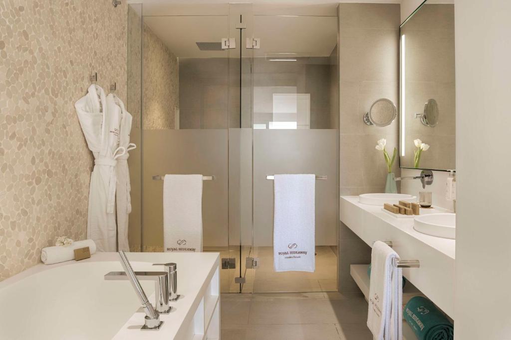 Mūsdienīgs vannas istabas interjers ar stikla dušas kabīni, baltām dvieļiem un elegantiem aksesuāriem, kas papildina relaksējošu atmosfēru.