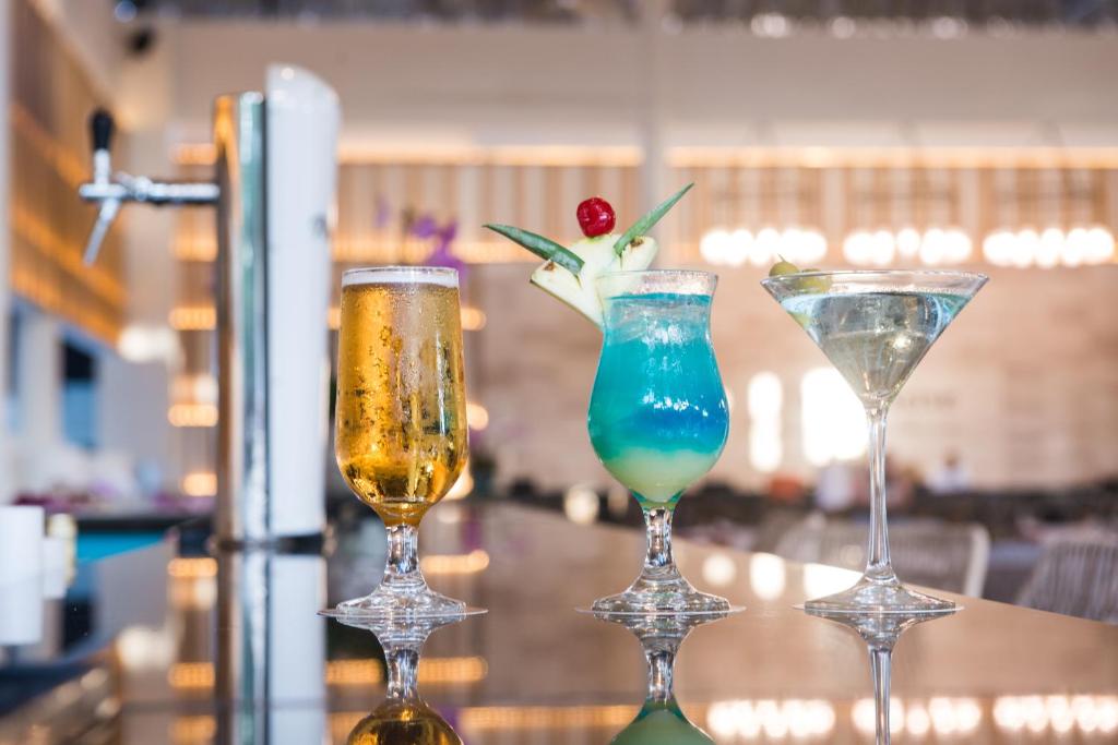 Trīs dzērieni, tostarp alus, spilgti zilais kokteilis ar ananāsu un ķiršu dekoru, un martini, novietoti uz bāra letes.