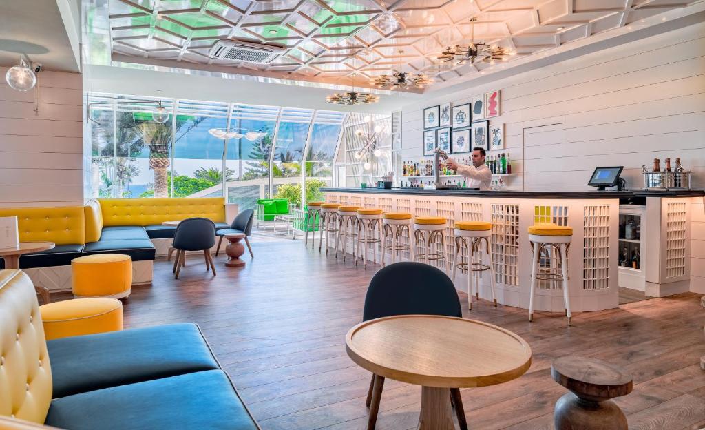 Mūsdienīgs bārs ar gaišu interjeru, kurā redzami dzelteni un zili krēsli, kā arī stilīga bārmeņa darbība. Liels logs sniedz skatu uz zaļo dabu ārpusē.