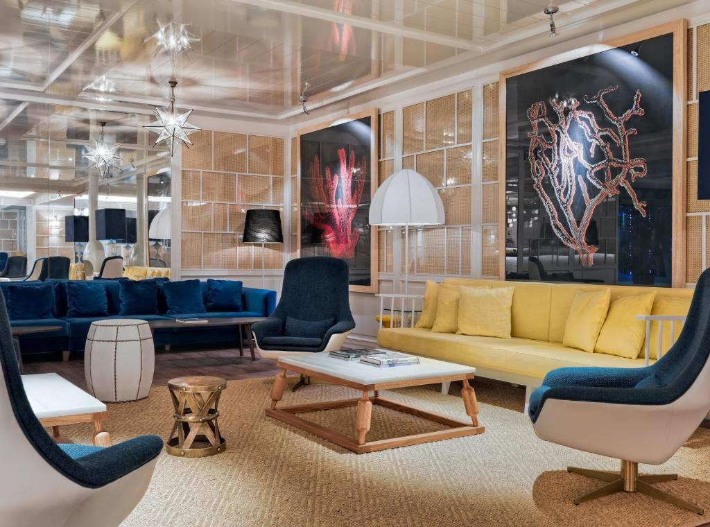 Mūsdienīgs viesību telpa ar ērtu dzeltenu dīvānu, zilo krēslu un mākslinieciskiem sienu attēliem, radot stilīgu un aicinošu atmosfēru.