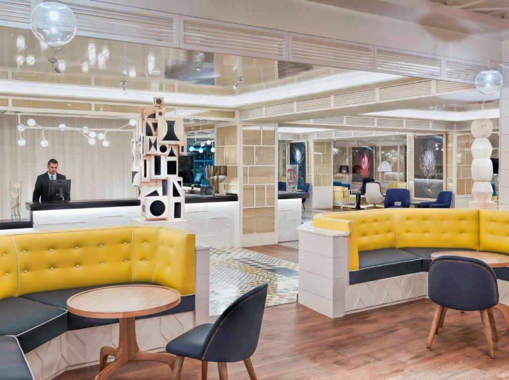 Mūsdienīgs viesnīcas reģistrācijas zonas interjers ar dzeltenām un melnām mēbelēm, kas apvieno funkcionālu dizainu un mākslinieciskas dekorācijas. Aiz letes stāv darbinieks, kamēr viesi atpūšas komfortablās sēdvietās.