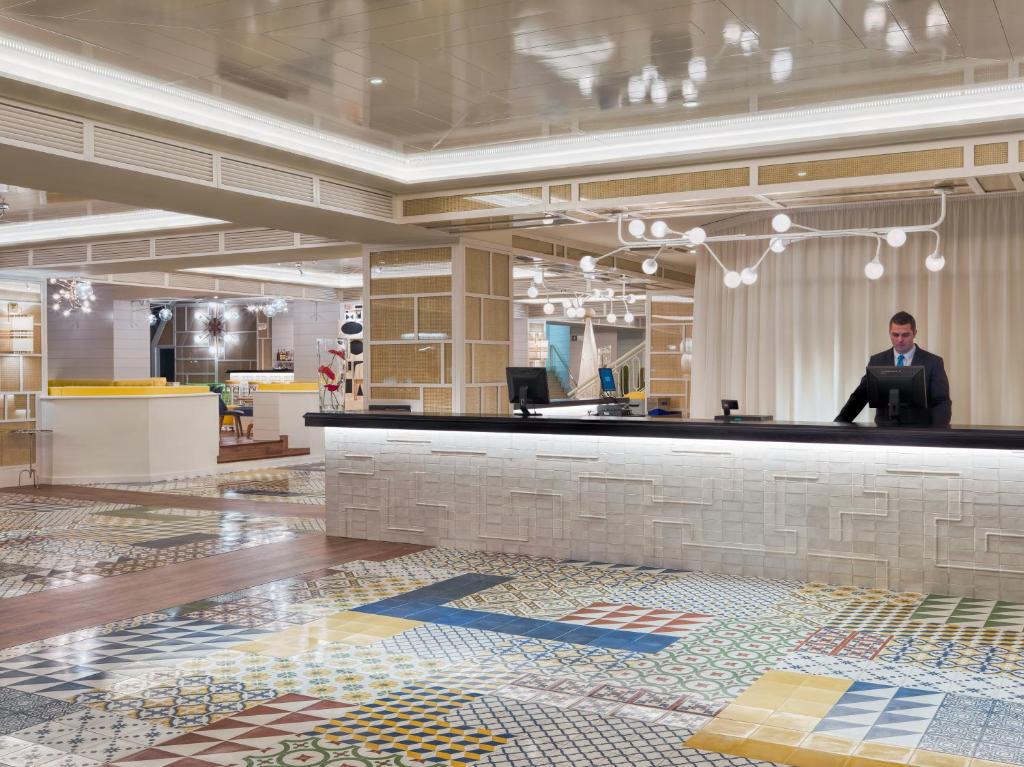 Mūsdienīga viesnīcas reģistratūras zona ar krāsainu flīžu grīdu un stilīgu apgaismojumu. Apskatāms viesnīcas darbinieks, kas apkalpo viesus.