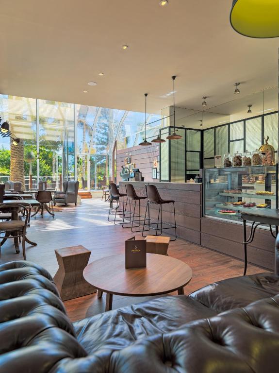 Mūsdienīgs kafejnīcas interjers ar plašām logu sienām, koka mēbelēm un siltu apgaismojumu, kas rada mājīgu atmosfēru. Kafejnīcā redzamas dažādas kūkas un dzērieni vitrīnā.