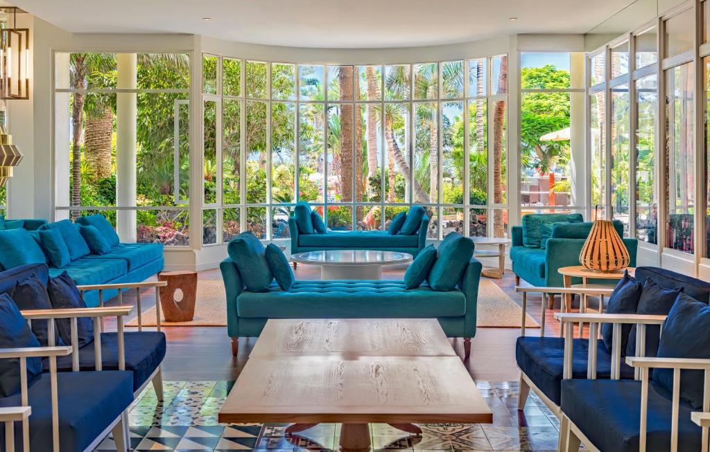 Mūsdienīgs un gaišs viesnīcas viesistabas interjers ar zilām dīvānām un logiem, caur kuriem redzams zaļš dārzs. Sēdvietas ir ērti izkārtotas ap centrālo galdu, radot relaksējošu atmosfēru.