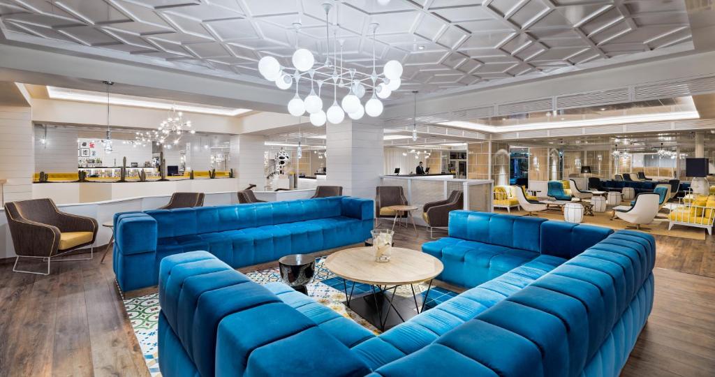 Mūsdienīgs viesnīcas uzgaidāmā telpas interjers ar spilgti zilu dīvānu, elegantu apgaismojumu un atpūtas zonām. Telpa ir plaša un gaiša, ar moderniem mēbeļu risinājumiem.