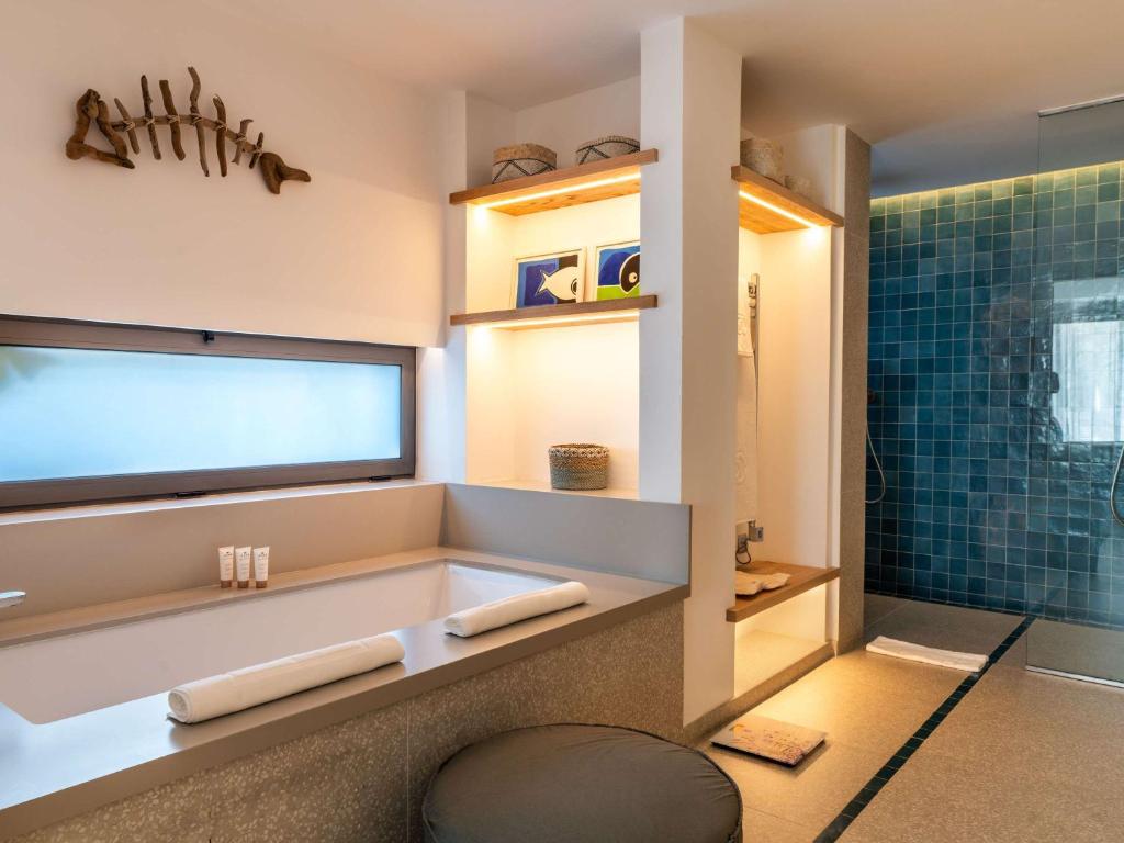 Mūsdienīgs vannas istabas interjers ar elegantu vannu, stilizētu apgaismojumu un flīžu sienām, radot relaksējošu atmosfēru. Uz plauktiem izvietoti dekoratīvi elementi un higiēnas piederumi.