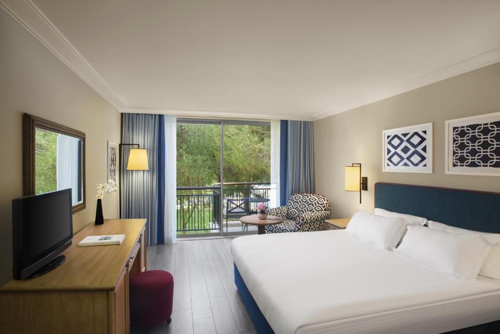 Mūsdienīgs viesnīcas numurs ar plašu logu, kas ved uz balkonu, ērti mēbelēts ar gultu, televizoru un atpūtas krēslu. Interjers ir gaišs un elegants, radot relaksējošu atmosfēru.