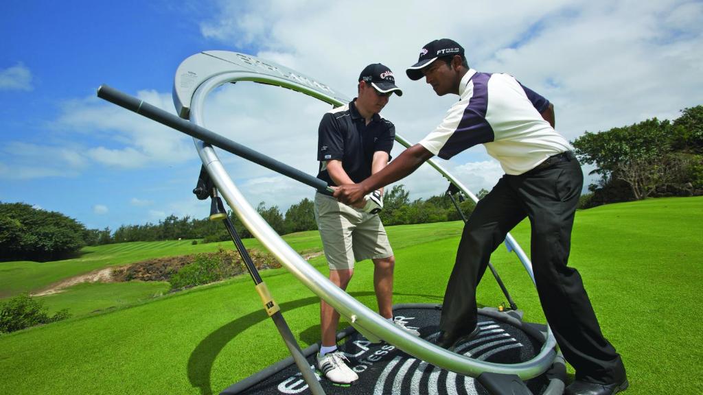 Divi golfa treneri strādā pie golfa treniņu aprīkojuma, lai uzlabotu spēlētāja tehniku. Fons ir zaļa golfa laukuma ainava ar ziliem debesīm.