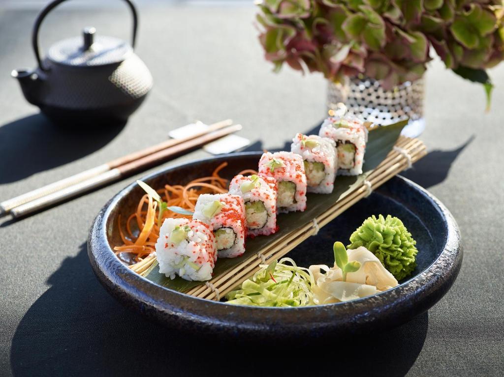 Sushi šķīvī ar garnelēm, wasabi un dārzeņu piedevām, dekorēts ar zaļām lapām un burkāniem. Blakus atrodas tējas kanna un kociņi.