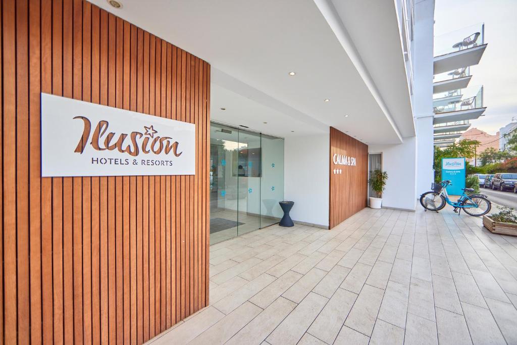 Ieeja viesnīcā ar zīmi "Illusion Hotels & Resorts" un "Calma & Spa", moderns dizains un koka apdare. Blakus redzamas velosipēdu stāvieta un zaļumi.
