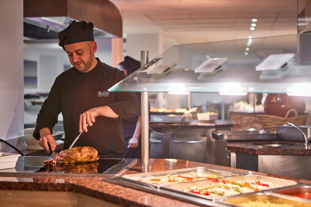 Šefpavārs, kas griež gaļu restorāna virtuvē, ar dažādiem ēdieniem un sastāvdaļām fonā.