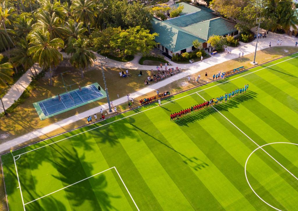 Futbola laukums ar divām komandām, kas stāv rindā, gatavojoties spēlei, apkārt palmām un skatītājiem. Ievērojams ainavas skats no augšas.