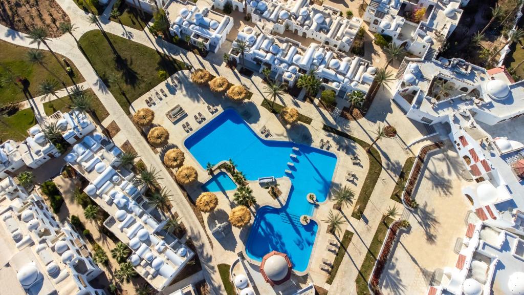 Apskatāma viesnīcas kompleksa ainava ar zilu baseinu un saulessargiem, kas atrodas apkārtne ar palmām un baltām ēkām. Ideāla vieta atpūtai un relaksācijai.