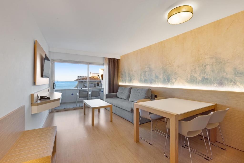 Mūsdienīgs viesnīcas numurs ar skatu uz jūru, iekārtots ar dīvānu, galdu un krēsliem. Telpa ir gaiša un eleganta, ar koka akcentiem un mūsdienīgu apgaismojumu.