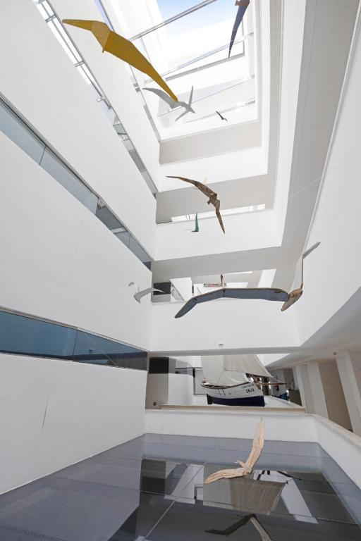 Mūsdienīga mākslas instalācija ar krāsainiem gaisa izstrādājumiem, kas karājas gaišā, baltā telpā ar stikla elementiem. Apakšā redzama laiva, kas papildina telpas estētiku.