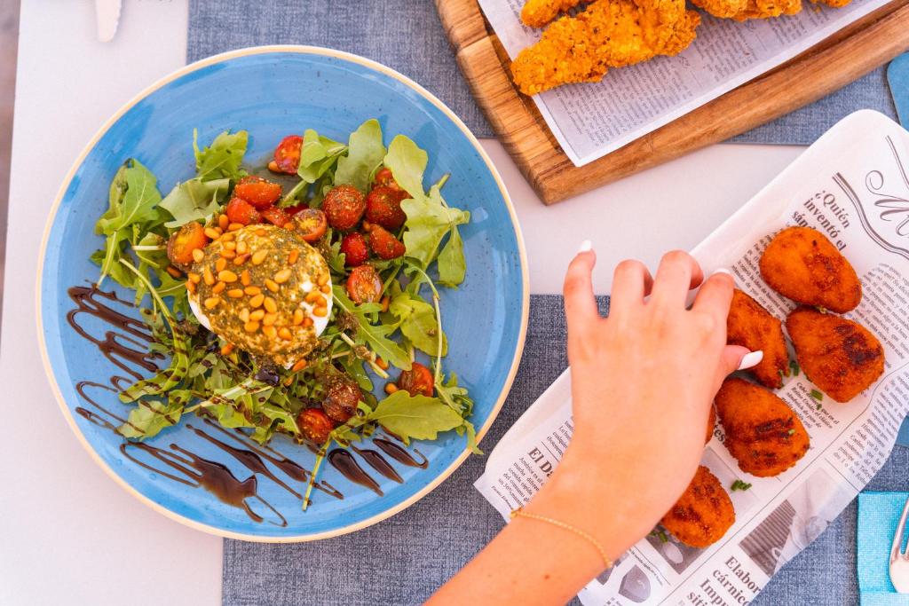 Svaigas salātu porcijas ar grauzdētiem tomātiem un riekstiem, kas pasniegti uz zila šķīvja, blakus ir cepti ēdieni uz papīra.