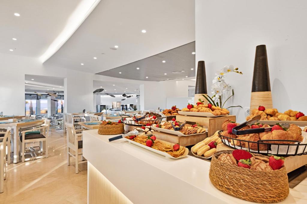 Brokastu bufete ar dažādiem konditorejas izstrādājumiem, augļiem un maizēm, kas pasniegta modernā restorānā ar gaišu interjeru. Eleganti galdi un krēsli papildina nesteidzīgu atmosfēru.
