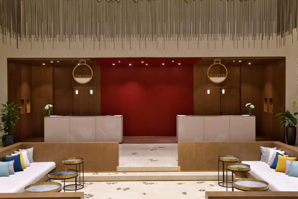 Modernas viesnīcas reģistratūra ar siltu apgaismojumu un elegantu dizainu, akcentēta ar sarkano sienu. Stilīgas sēdvietas un augi piešķir telpai viesmīlību.
