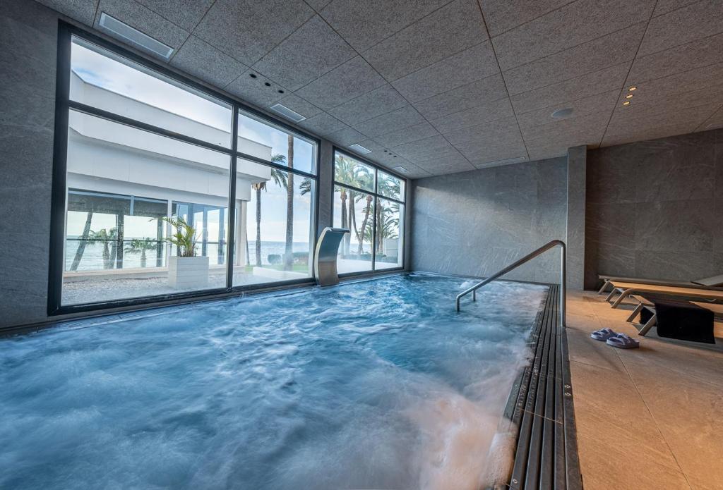 Mūsdienīgs spa ar iekšējo baseinu, caur kuru redzama pludmale un palmas. Ūdens ir viļņains, un telpā ir daudz dabiskās gaismas.
