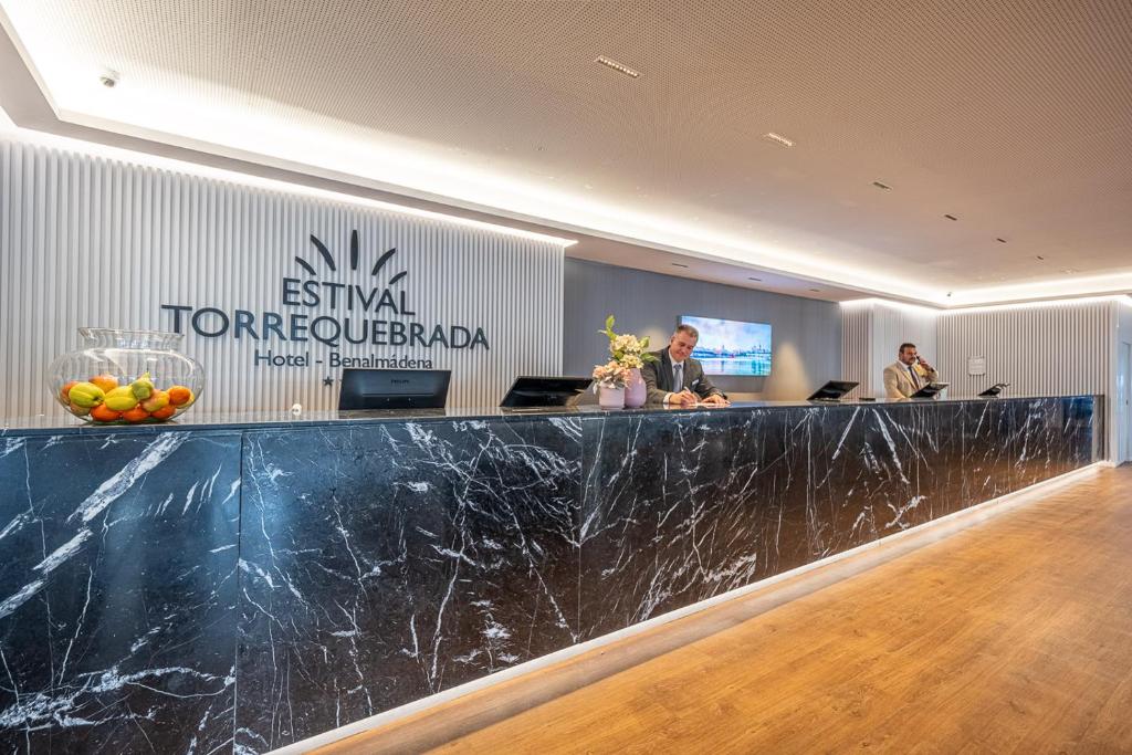 Recepcija viesnīcā Estival Torrequebrada, Benalmadena, ar modernu dizainu un draudzīgu personālu. Uz letes atrodas augļu trauks un ziedu kompozīcija.