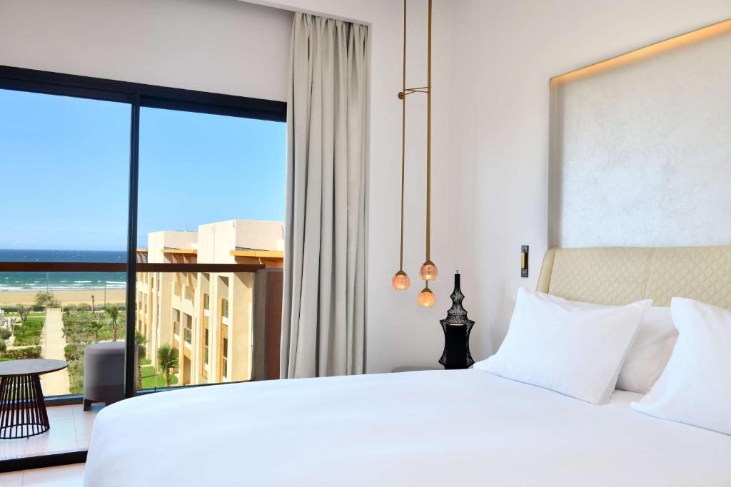 Moderns viesnīcas numurs ar gultu, elegantu apgaismojumu un skatu uz jūru. Logā redzama ainava ar pludmali un dārzu.